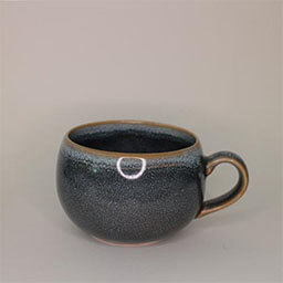 A small round cup with a handle facing right 
                            A dark blue cup with a shiny stoneware glaze 
                            An empty cup in studio